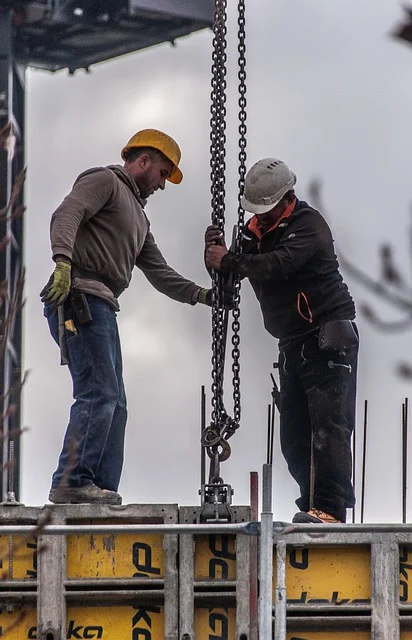 Deux ouvriers sur un chantier de construction ajustant une chaîne de levage, portant des casques de sécurité et des équipements de protection.