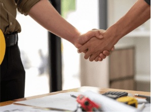 Deux professionnels se serrant la main, symbolisant un accord ou un partenariat, avec un casque de chantier jaune visible à la ceinture et des documents techniques sur la table